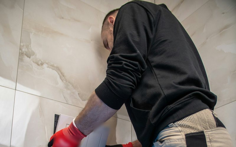 Plumber in a black hoodie fitting a flush plate on a tiled bathroom wall